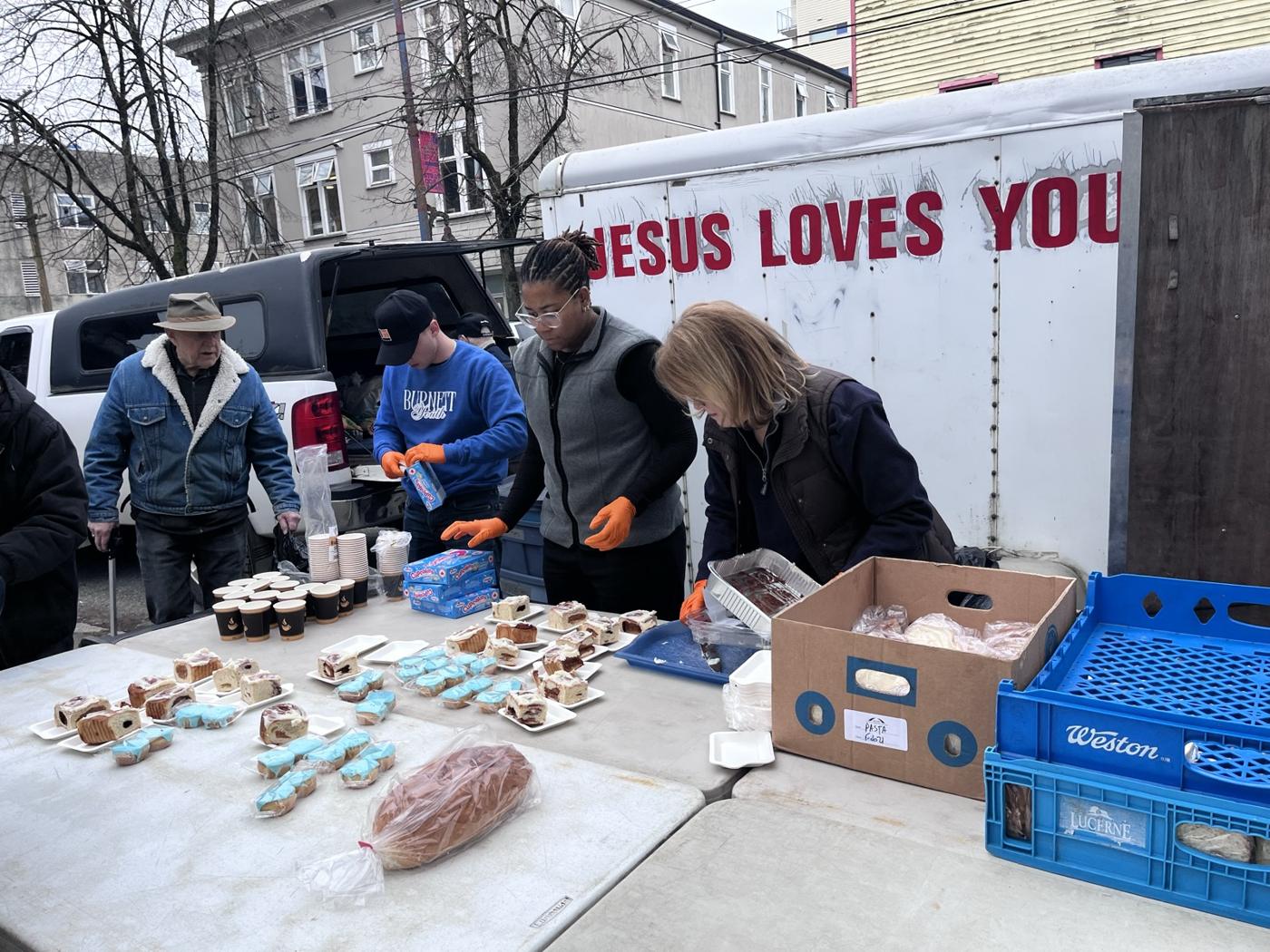 Volunteers handing out food from folding tables beside the Jesus Loves You trailer